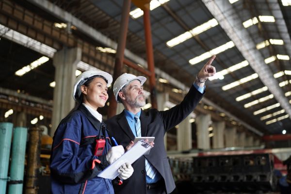 A male executive pointing out structural details to a female industrial worker with a walkie-talkie and clipboard during a plant inspection