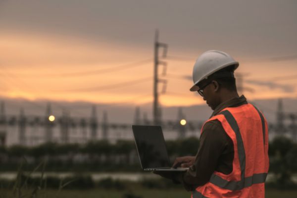 An engineer wearing a white hard hat and high-vis vest using a laptop outdoors near power lines at sunset