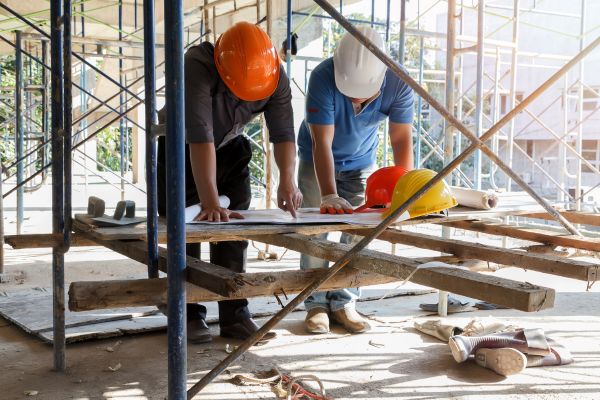 Two construction workers in hard hats leaning over a wooden scaffold workbench to review architectural plans