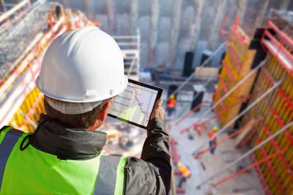 Construction inspector viewing digital plans and progress metrics on a tablet while overlooking a large-scale building excavation site