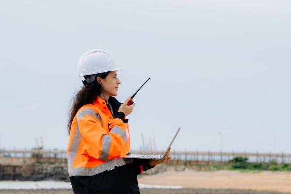 Female construction engineer in a white hard hat holding a laptop and talking on a walkie-talkie outdoors